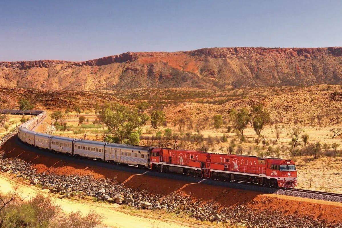 The Ghan mit roten Lokomotiven in einer weiten Kurve vor einer roten Hügelkette im australischen Outback